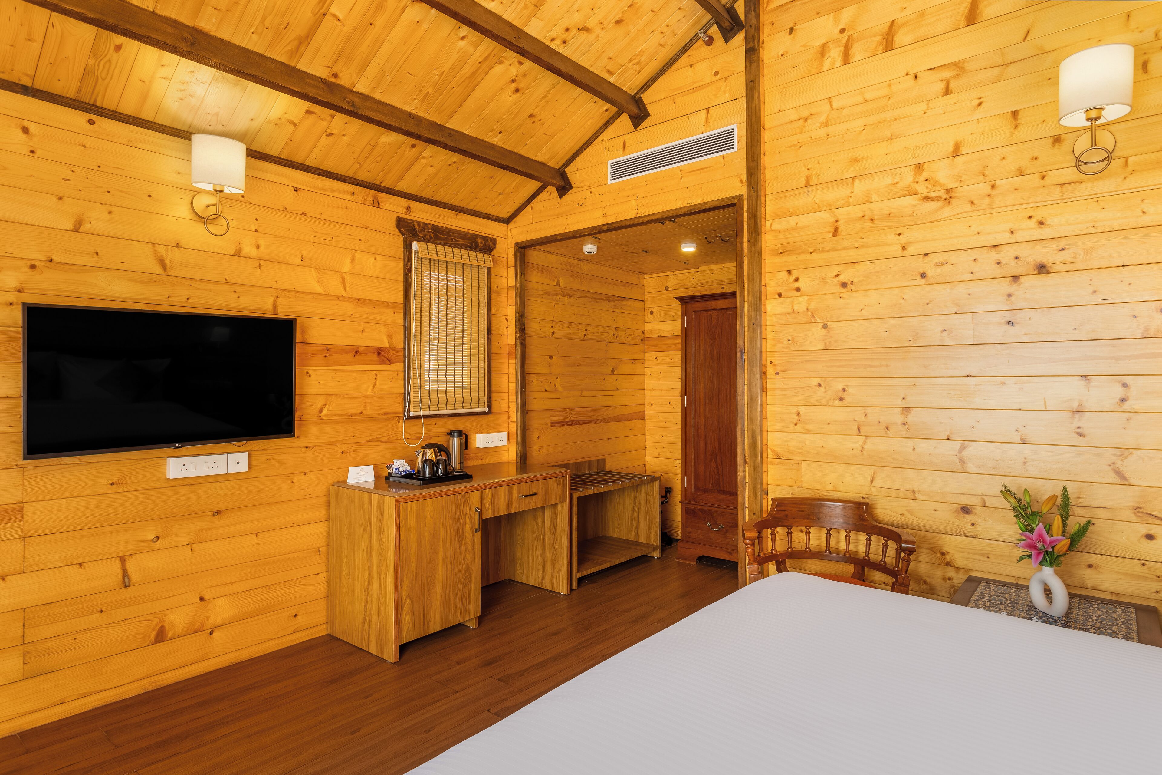 A wooden cabin interior with a bed, a desk, a chair, a TV, and a wall-mounted lamp.