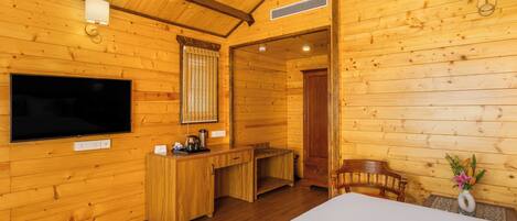 A wooden cabin interior with a bed, a desk, a chair, a TV, and a wall-mounted lamp.
