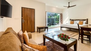 A modern hotel room with a sofa, a coffee table with flowers, a bed with a headboard, a desk, and a television. - Bogmallo Beach Resort (Marmagao)