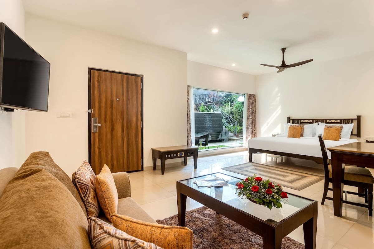 A modern hotel room with a sofa, a coffee table with flowers, a bed with a headboard, a desk, and a television.