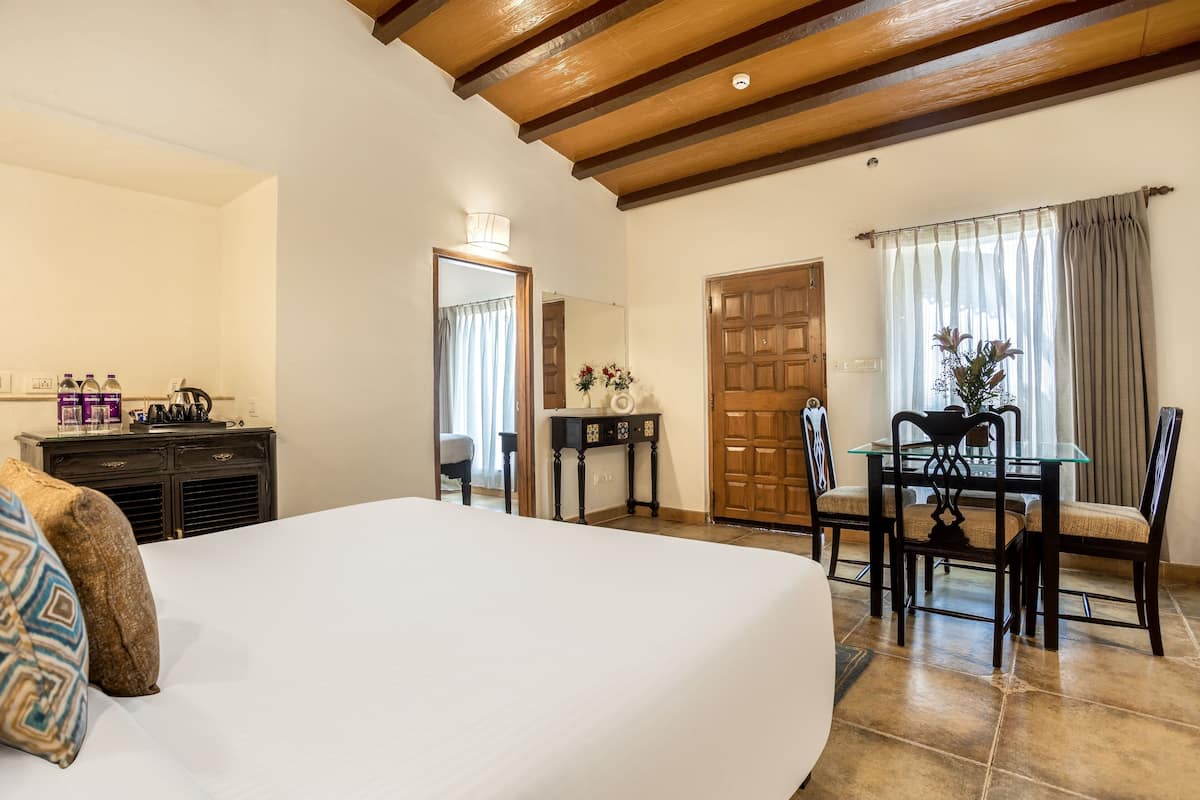 A hotel room with a large bed, a wooden ceiling, a dining table with chairs, a small table with a vase, and a door.