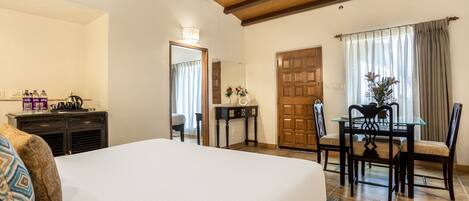 A hotel room with a large bed, a wooden ceiling, a dining table with chairs, a small table with a vase, and a door.