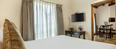 Habitación de hotel con una cama grande, un televisor en la pared, un escritorio con lámpara y un área de comedor visible a través de una puerta abierta.