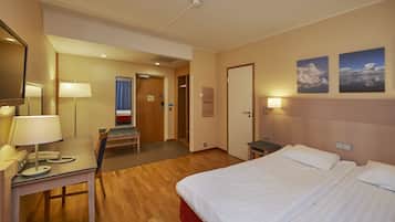 Quarto de hotel com cama, escrivaninha, cadeira e uma pequena mesa com abajur. Há uma porta que dá para um banheiro e um guarda-roupa.
