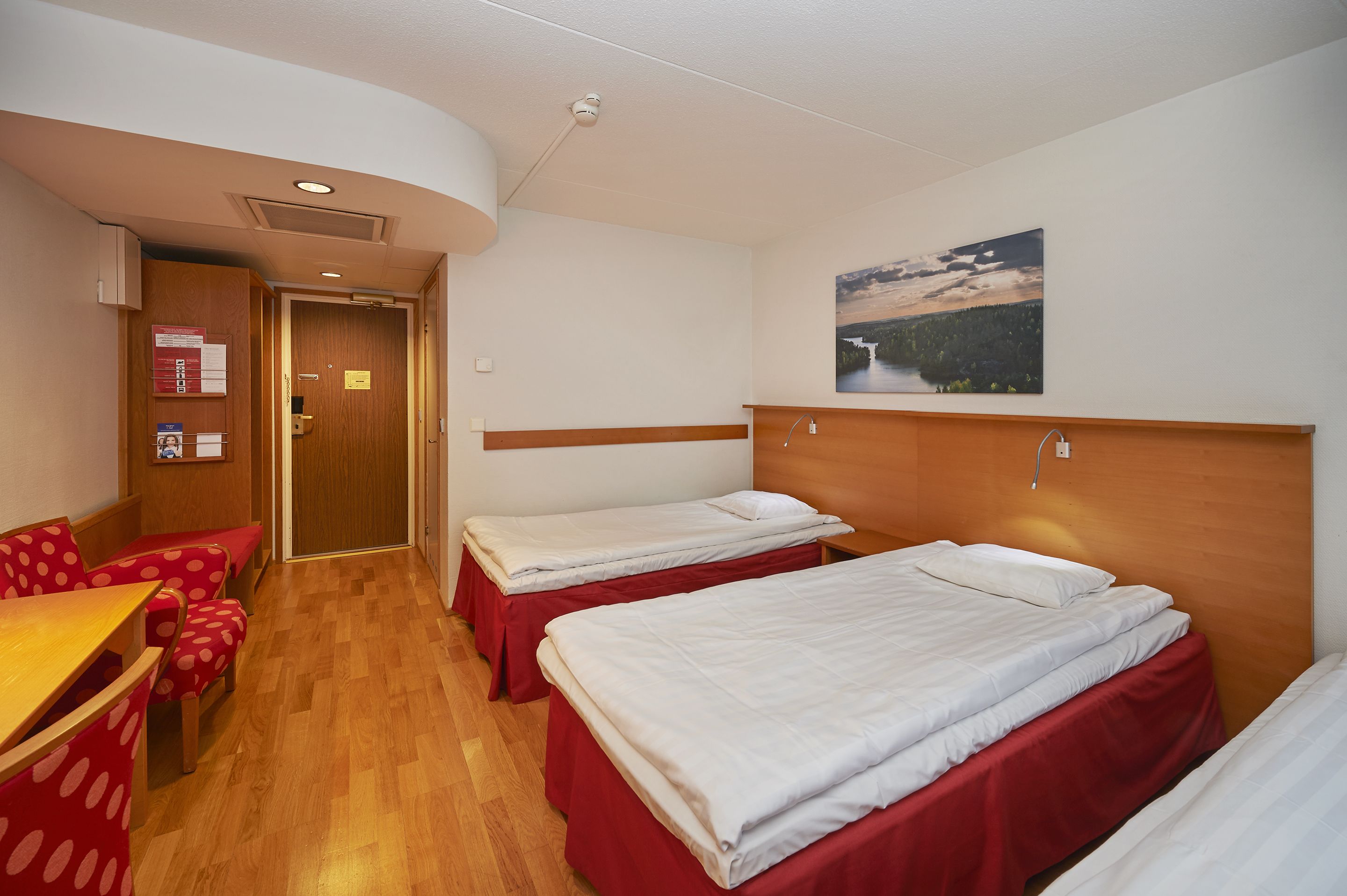 Une chambre d’hôtel avec deux lits simples, une tête de lit en bois, une table en bois avec des chaises rouges et un tableau au mur.