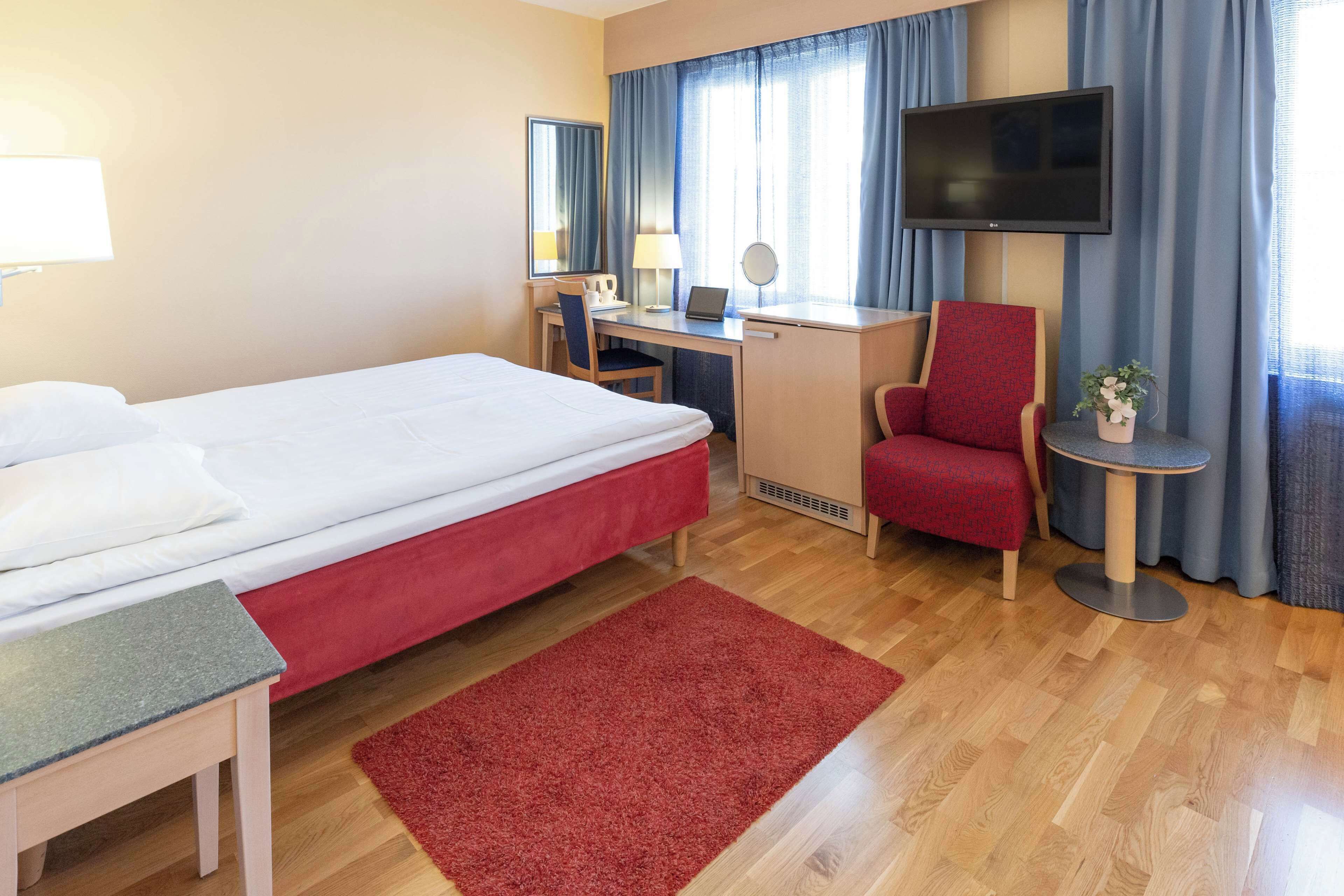 A hotel room with a bed, desk, chair, TV, and a small table with a plant.