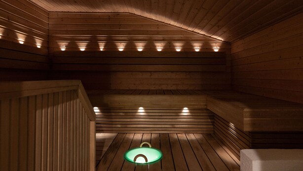 Una sauna con paredes y bancos de madera, un elemento central iluminado en verde y un banco a la derecha.