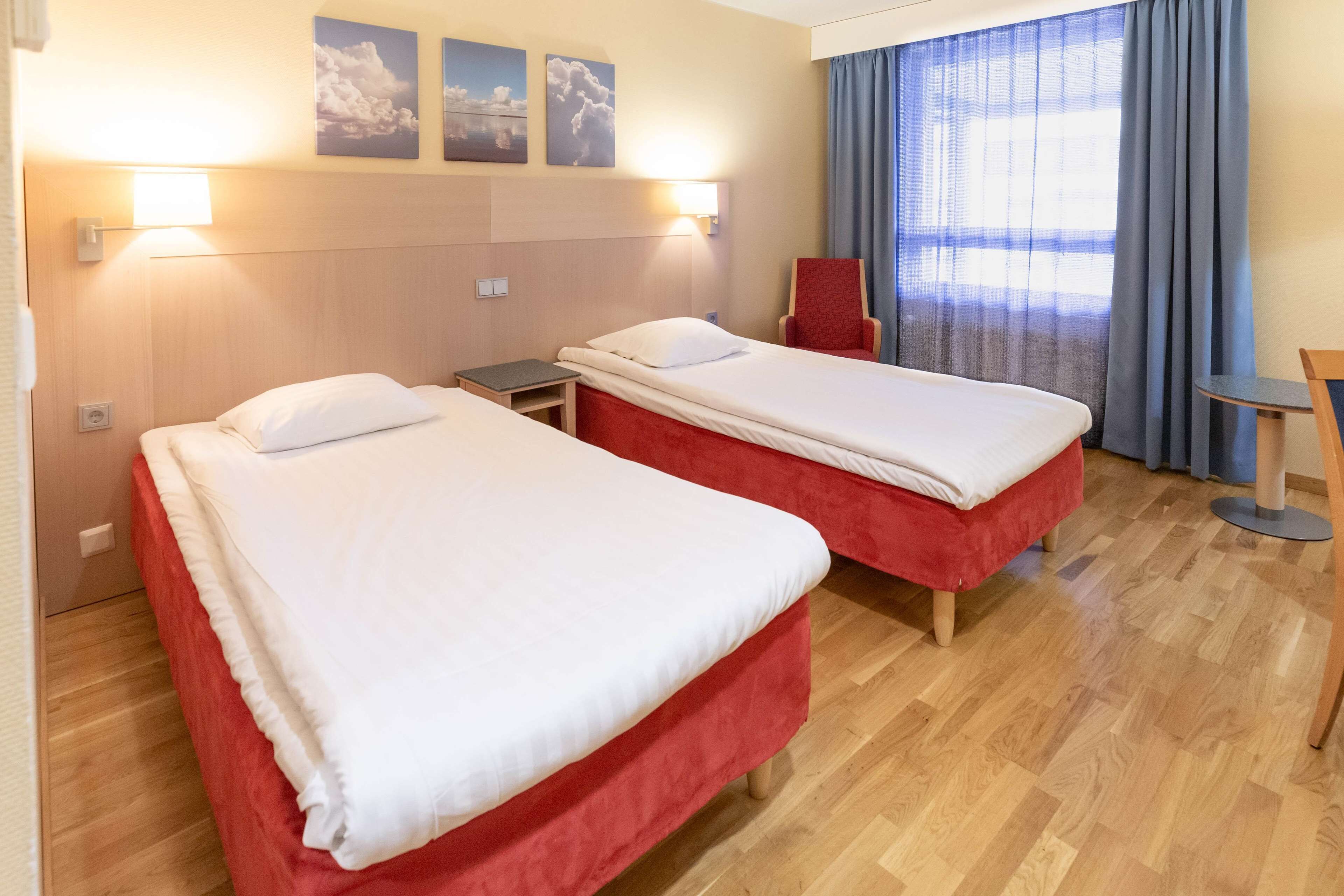 A hotel room with two single beds, a small table, a chair, and a window with curtains.