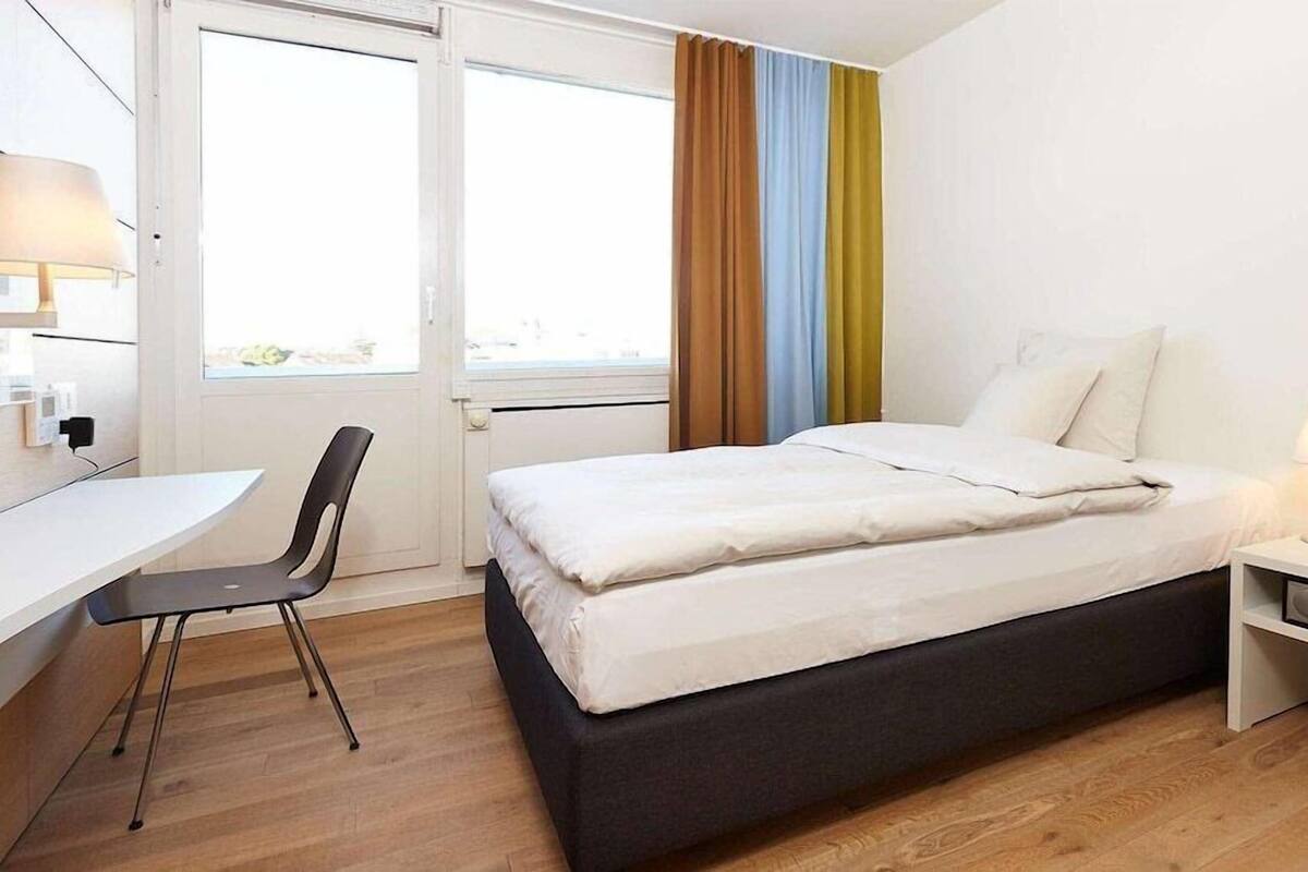 Une chambre d’hôtel avec un lit, une table de chevet, une lampe, une chaise, un bureau et une vue par la fenêtre.