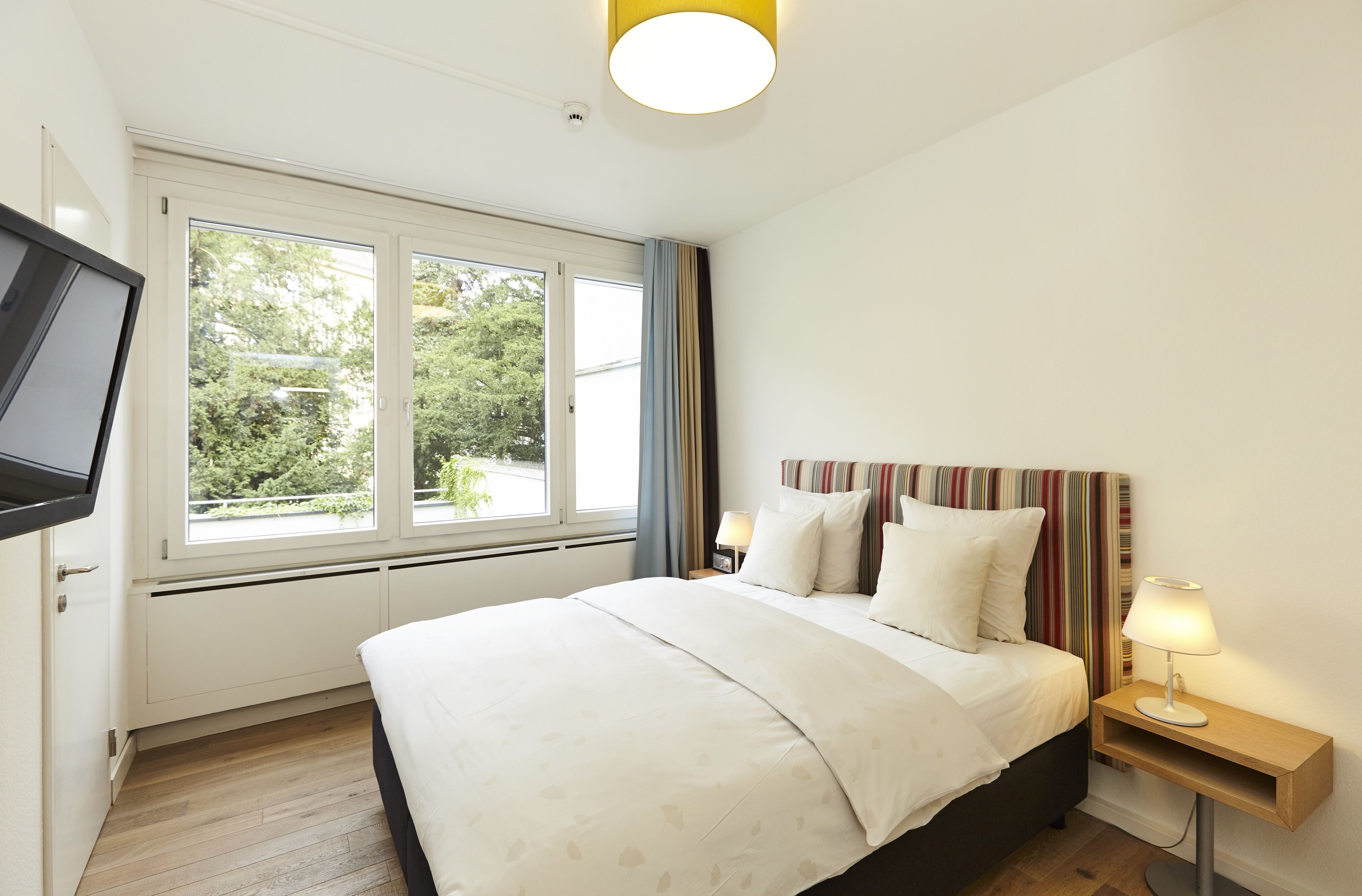 Een slaapkamer met een bed, een nachtkastje met een lamp, een aan de muur bevestigde televisie en een raam met gordijnen.
