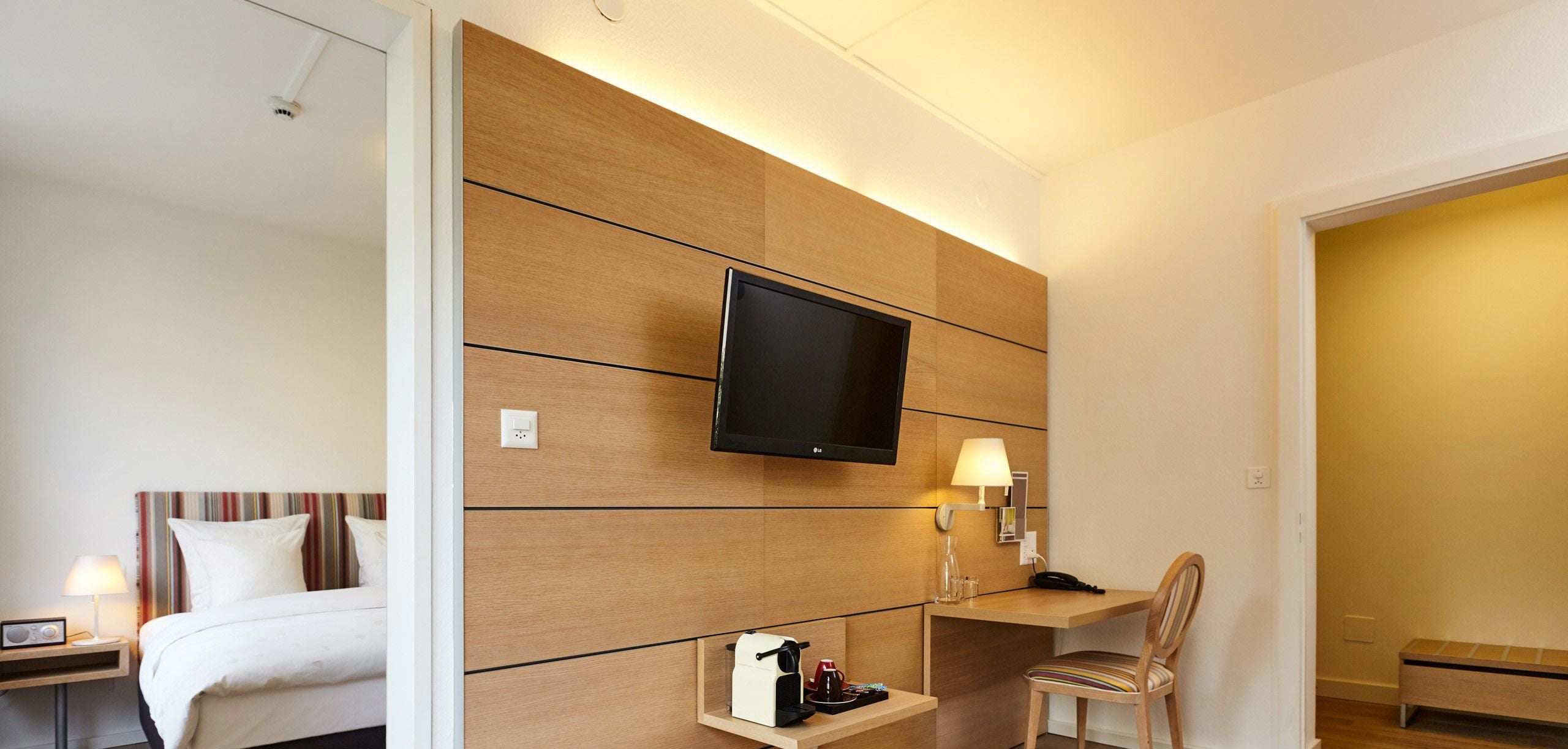 A hotel room with a bed, a desk with a chair, a television mounted on the wall, and a bedside table with a lamp and a radio.