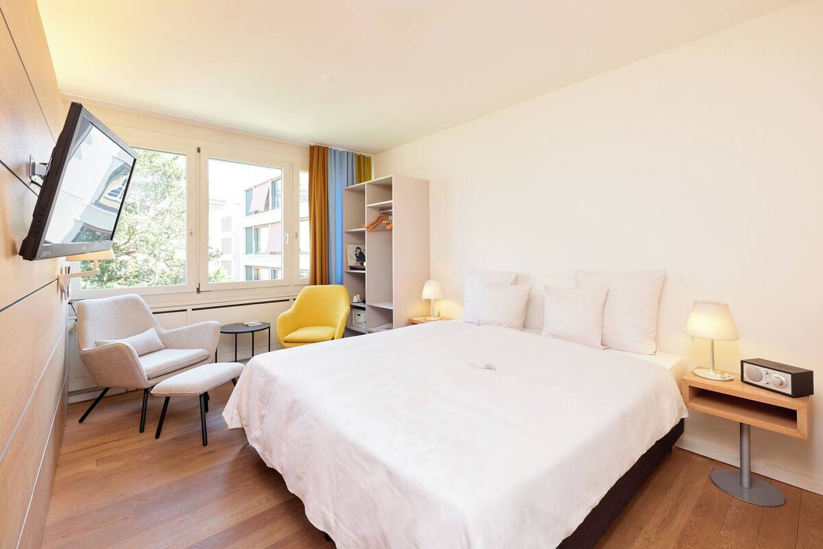 Une chambre d’hôtel moderne dotée d’un grand lit, d’un bureau avec une chaise, d’un téléviseur fixé au mur et d’une fenêtre donnant sur des immeubles.