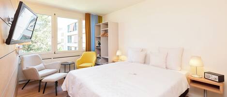 A modern hotel room with a large bed, a desk with a chair, a TV mounted on the wall, and a window with a view of buildings.