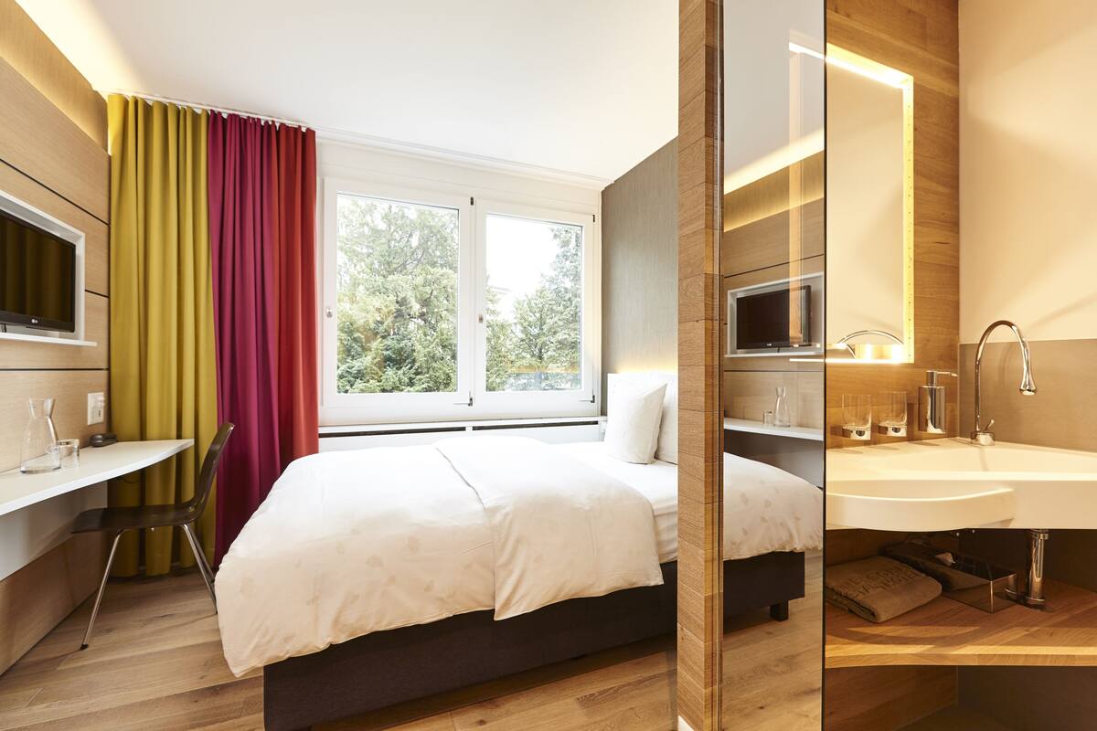 A modern hotel room with a bed, a desk, a TV, a large window with curtains, and a bathroom with a sink and mirror.