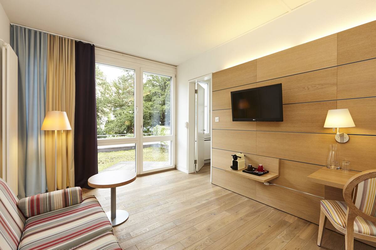 A modern hotel room with a sofa, a small round table, a TV mounted on a wooden panel wall, and a sliding glass door leading to an outdoor area.