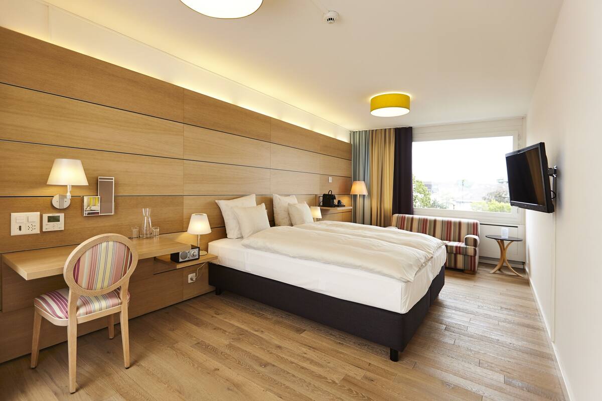 A modern hotel room with a large bed, a desk, a chair, a TV, and a window with a view.