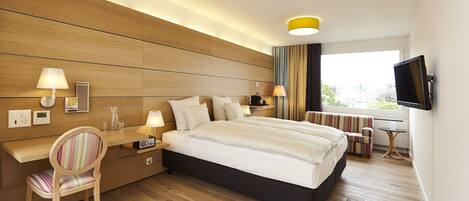 A modern hotel room with a large bed, a desk, a chair, a TV, and a window with a view.