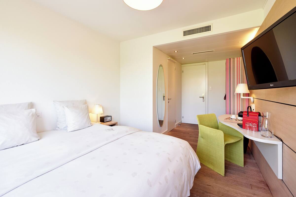 A hotel room with a bed, a desk with a TV, a chair, and a mirror.