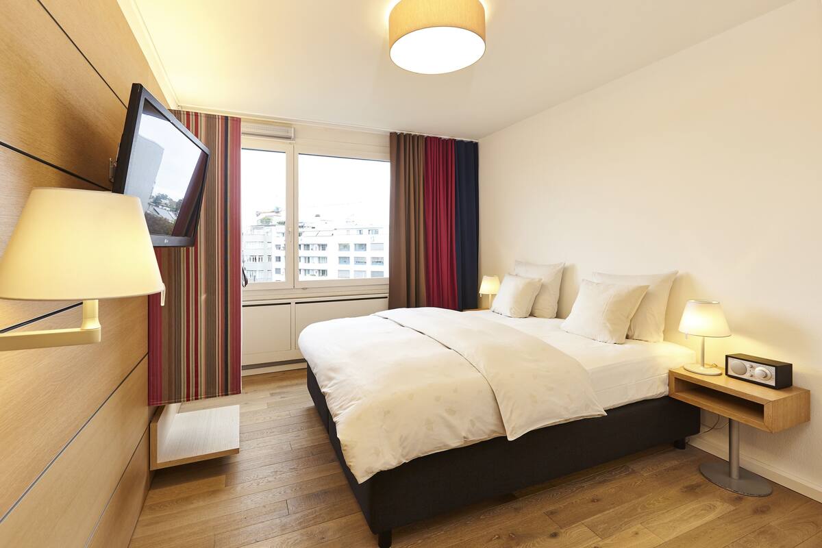 A hotel room with a bed, bedside lamps, a TV mounted on the wall, and a window with curtains.