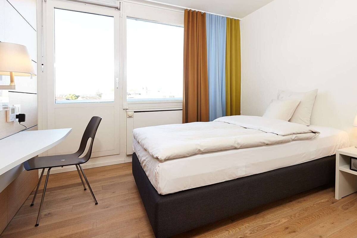 Une chambre d’hôtel moderne équipée d’un lit, d’une table de chevet, d’une lampe et d’un bureau avec une chaise.