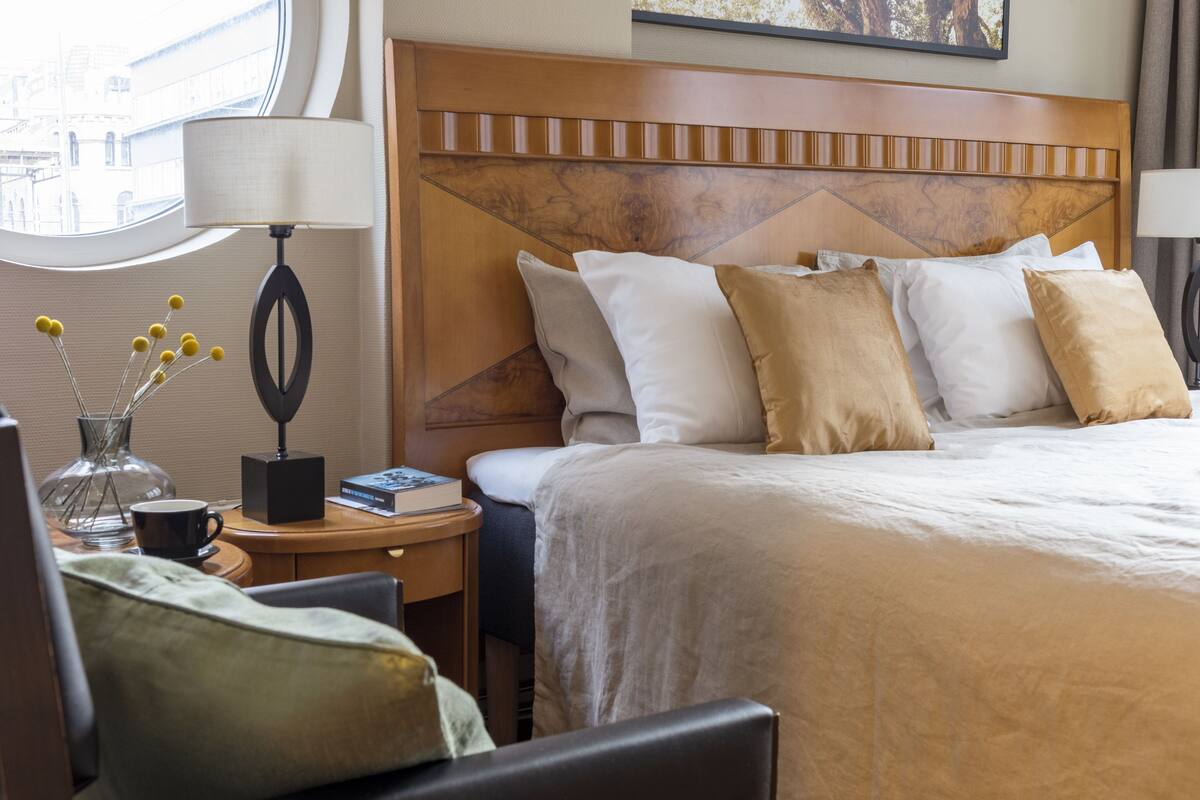 A hotel room with a large bed, a wooden headboard, a bedside table with a lamp, a vase with yellow flowers, and a window with a view of buildings.