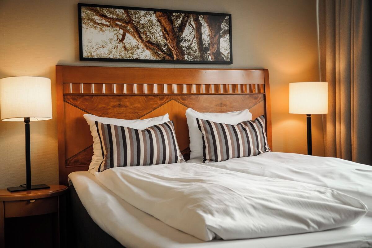 A hotel room with a large bed, two bedside lamps, a wooden headboard, and a framed picture on the wall.