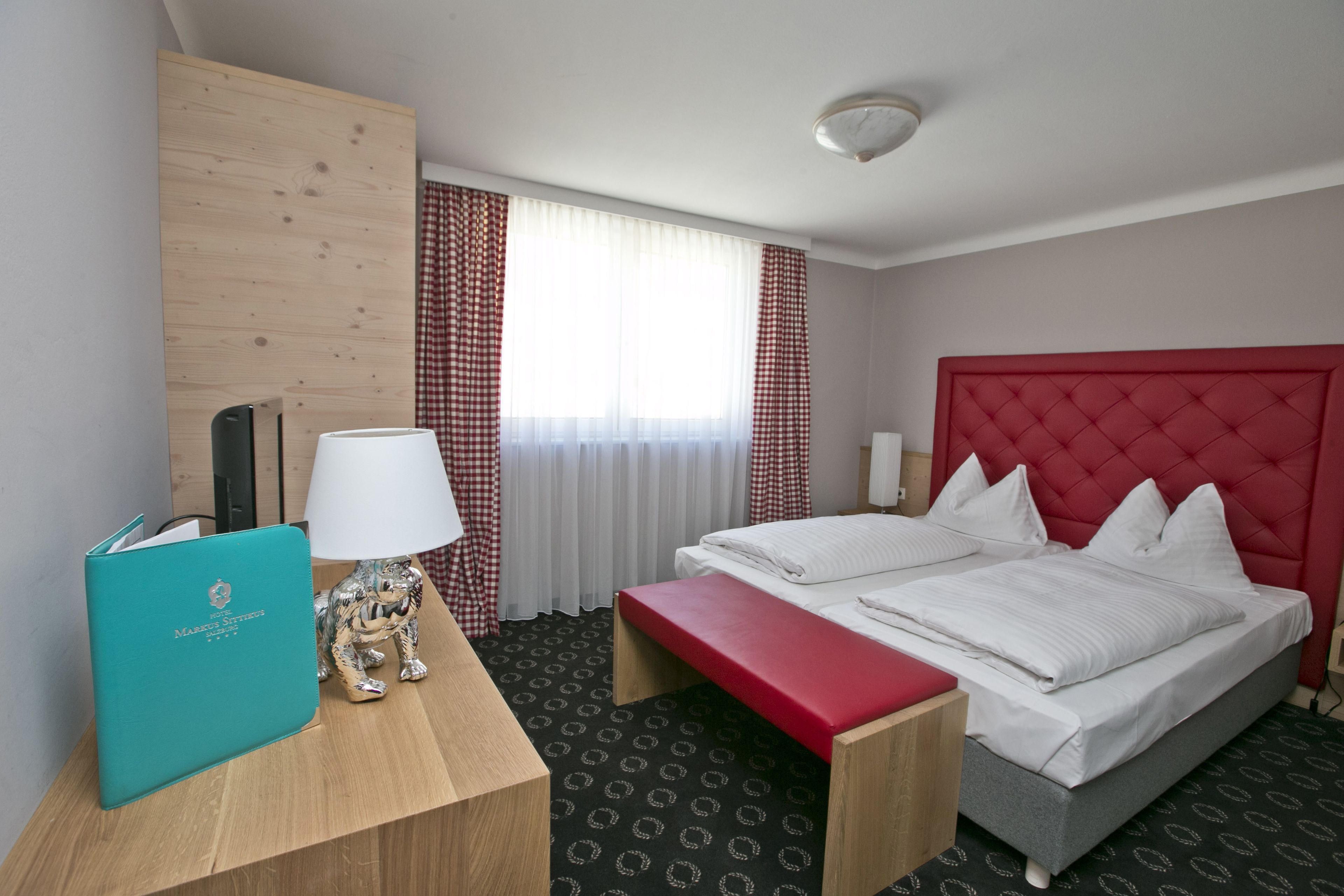 A hotel room with a bed, a desk with a lamp, a book, and a red bench.