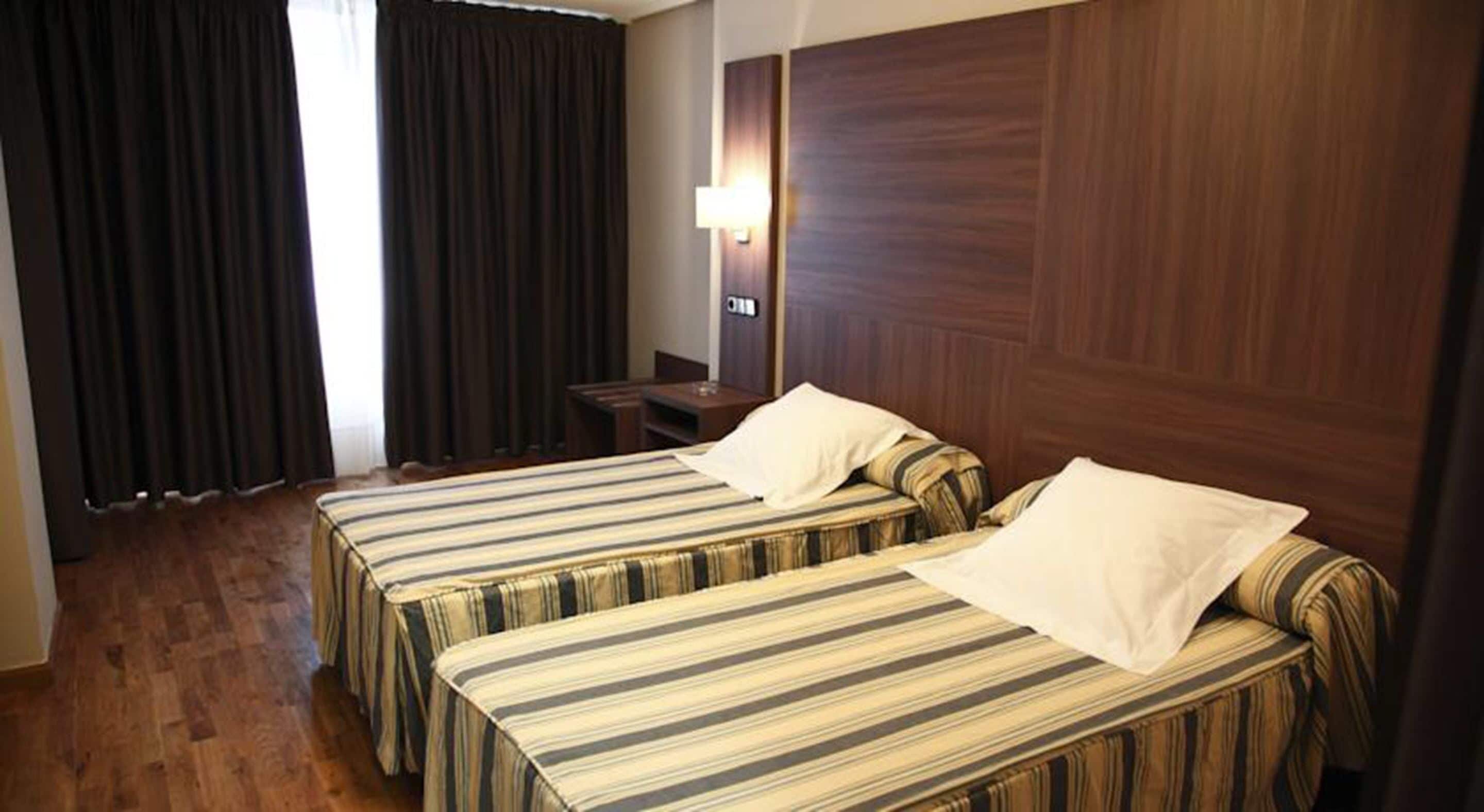 A hotel room with two beds, wooden flooring, a nightstand, a lamp, and dark curtains.
