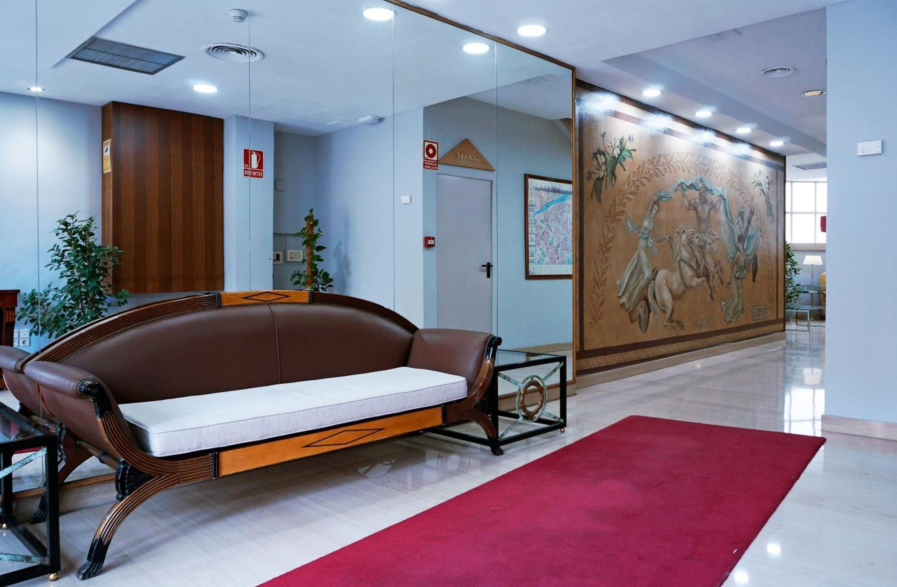 Un vestíbulo con un sofá de cuero marrón, una alfombra roja, una mesa de centro de vidrio y un mural en la pared.