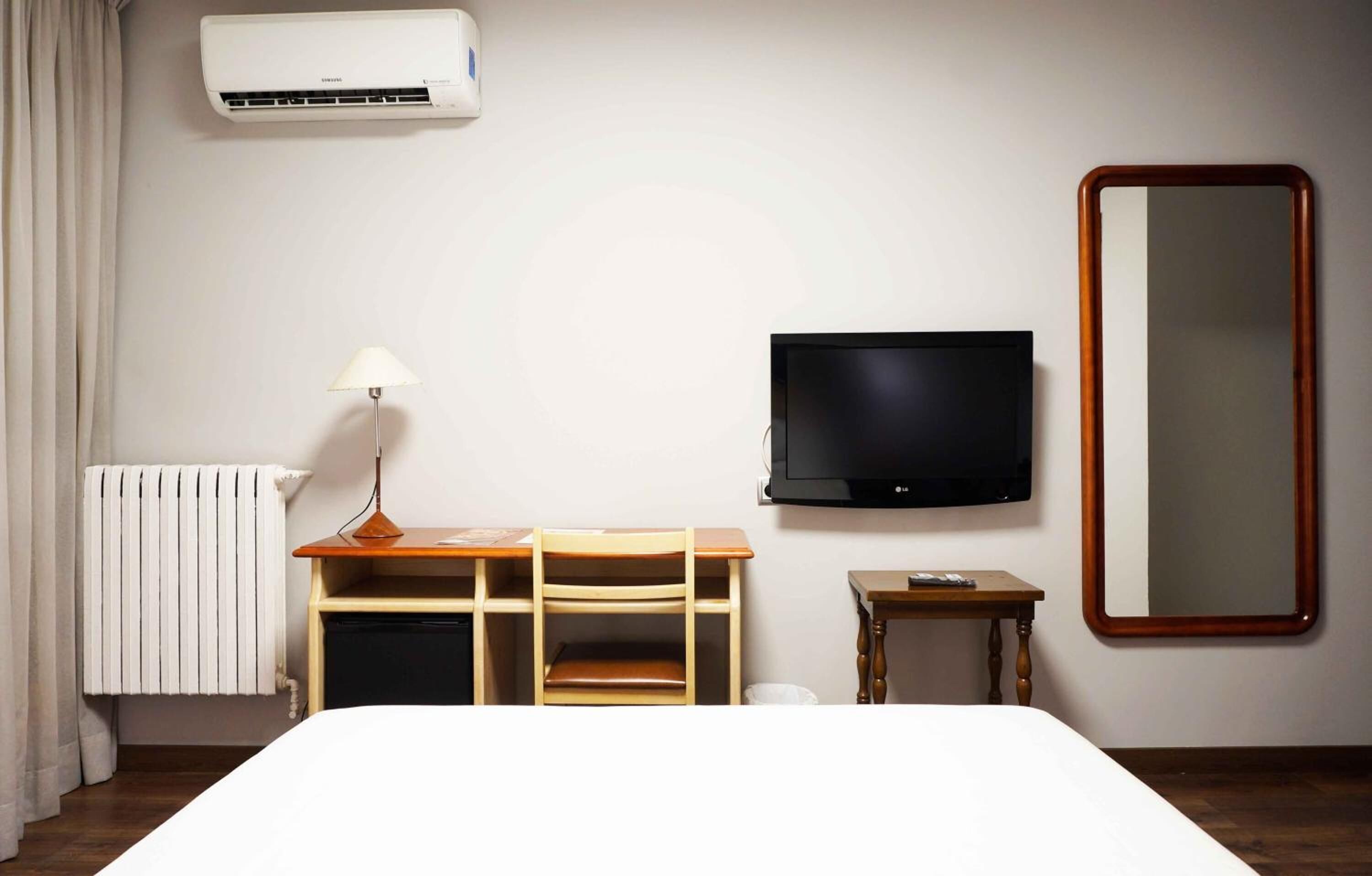 A hotel room with a bed, a desk with a lamp, a TV, a mirror, and an air conditioner.