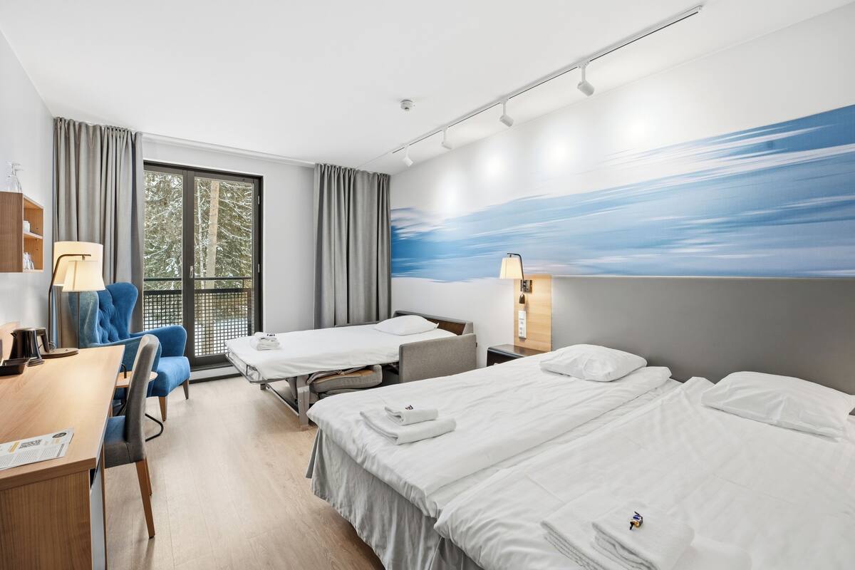A hotel room with two beds, a desk, a chair, a window with curtains, and a wall mural.