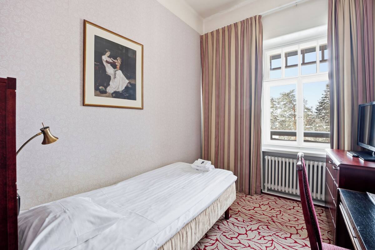 A hotel room with a bed, a framed picture on the wall, a window with curtains, a radiator, and a desk with a television.