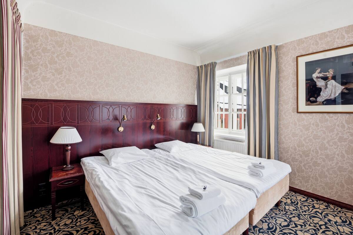 A hotel room with a large bed, two bedside lamps, a wooden headboard, a patterned carpet, and a painting on the wall.