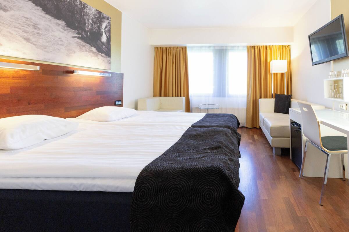 A hotel room with a large bed, a desk with a laptop, a chair, a TV, and a window with curtains.