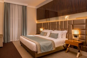 A hotel room with a large bed, a wooden headboard, bedside lamps, and a view of the city through the window.