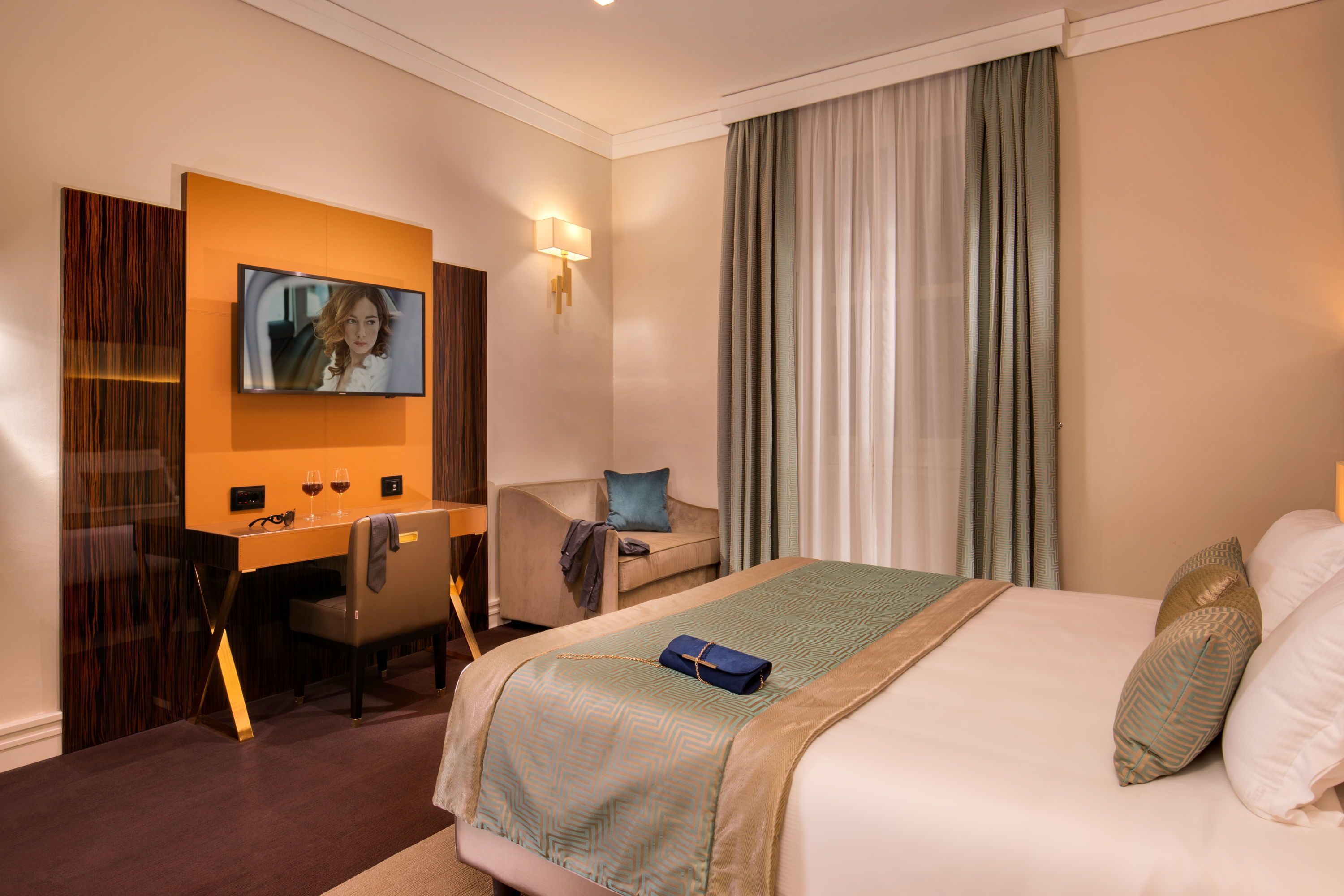 A hotel room with a bed, a desk with a television, a chair, and a window with curtains.