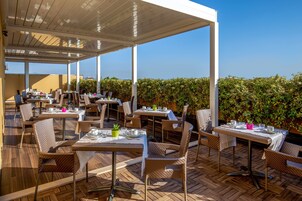 An outdoor dining area with tables and chairs, covered by a roof, and surrounded by greenery.