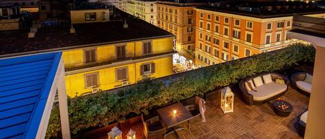 A rooftop terrace with outdoor furniture, a fire pit, and a view of the cityscape at dusk.