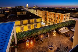 A rooftop terrace with outdoor furniture, a fire pit, and a view of the cityscape at dusk.