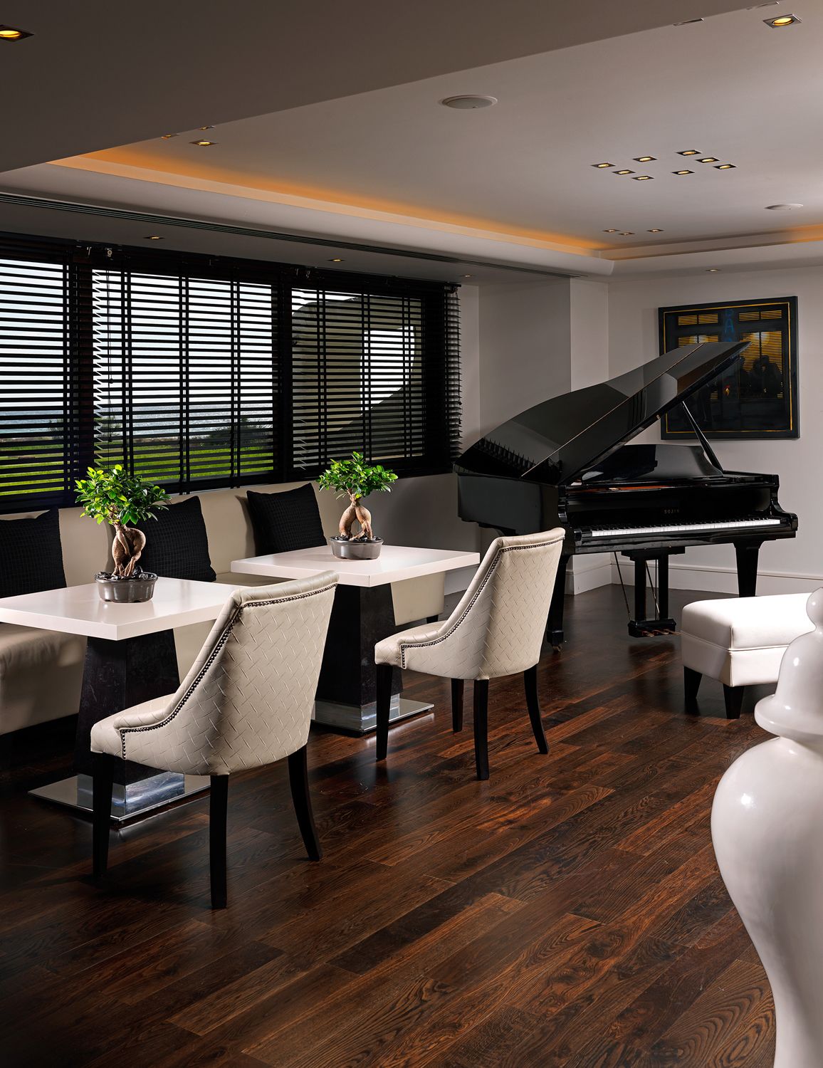 A modern living room with a grand piano, a dining area with white furniture, and large windows with blinds.