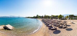 A beach with sun loungers and umbrellas, a clear view of the sea, and a bridge in the background. - Porto Rio Hotel Casino (Patras)