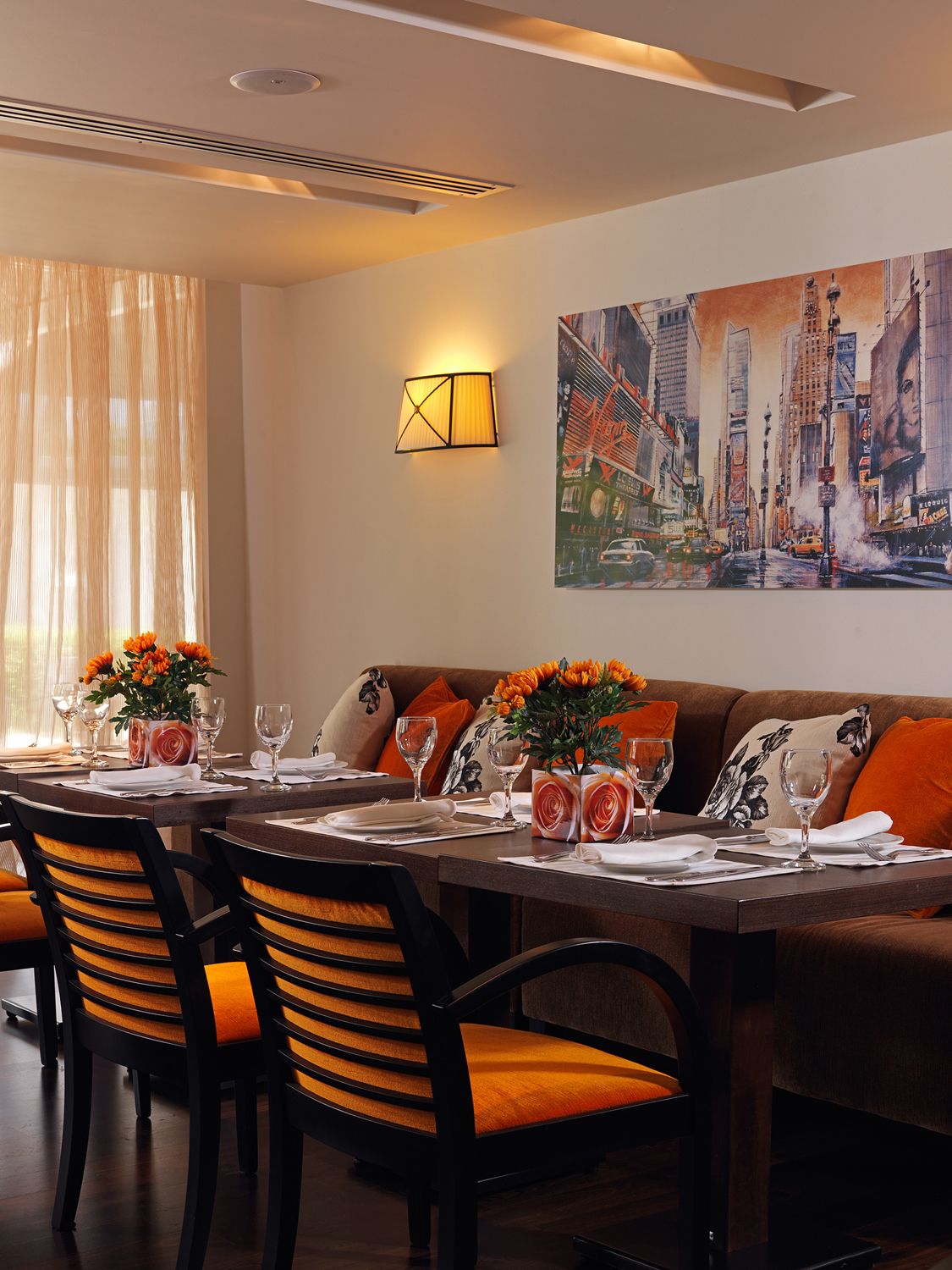 Un comedor con una mesa dispuesta para dos, un sofá con cojines naranjas y un cuadro de un paisaje urbano en la pared.