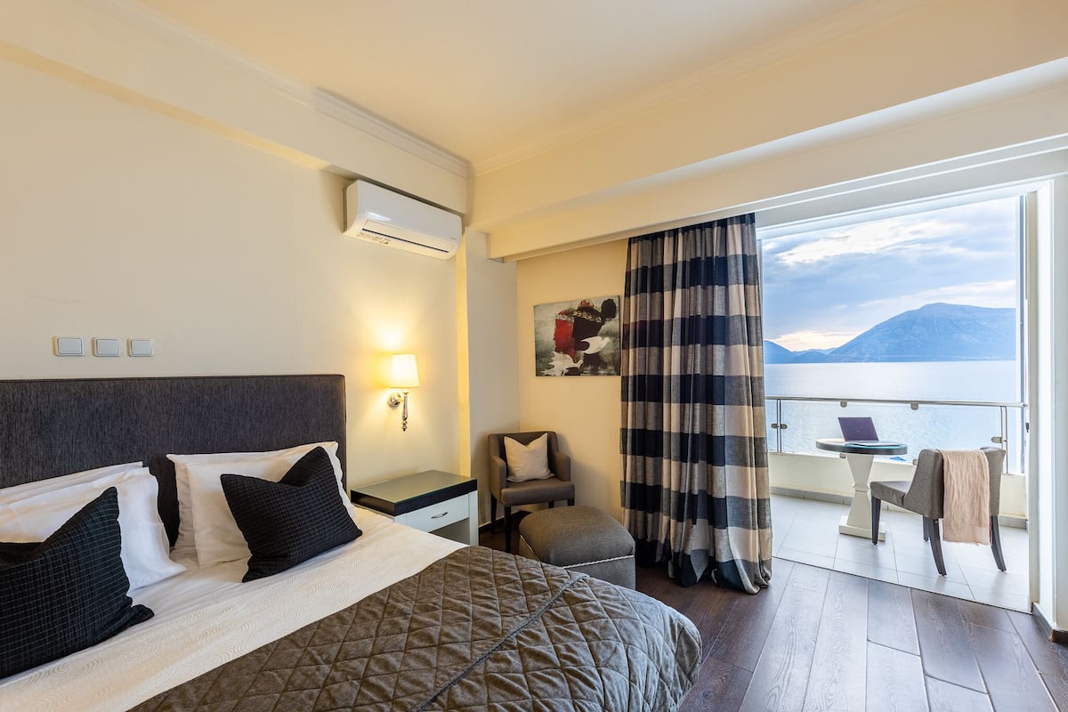 A hotel room with a bed, a desk, a chair, and a balcony with a view of the sea and mountains.