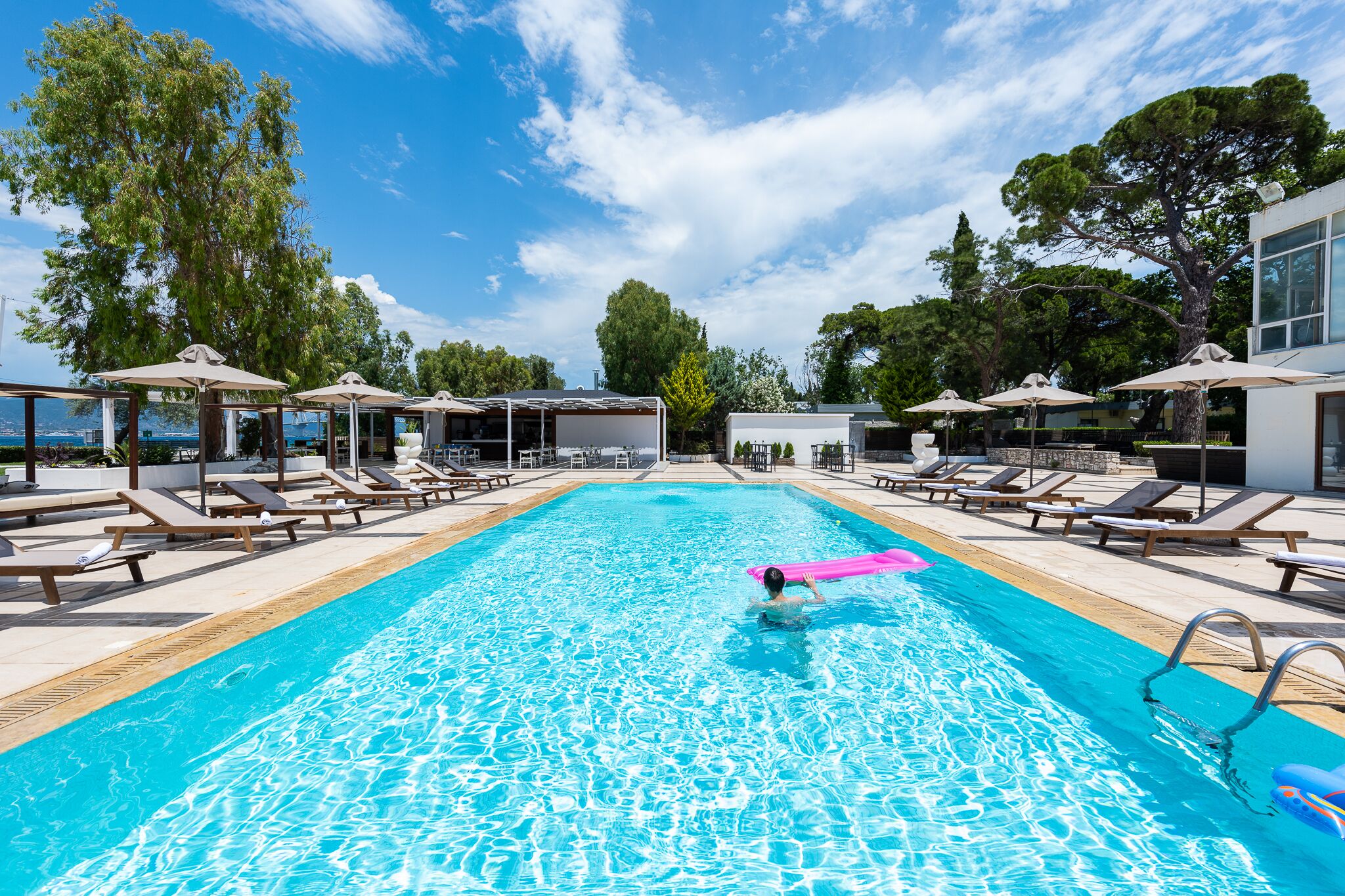 A swimming pool with lounge chairs and umbrellas, surrounded by trees and a clear sky.