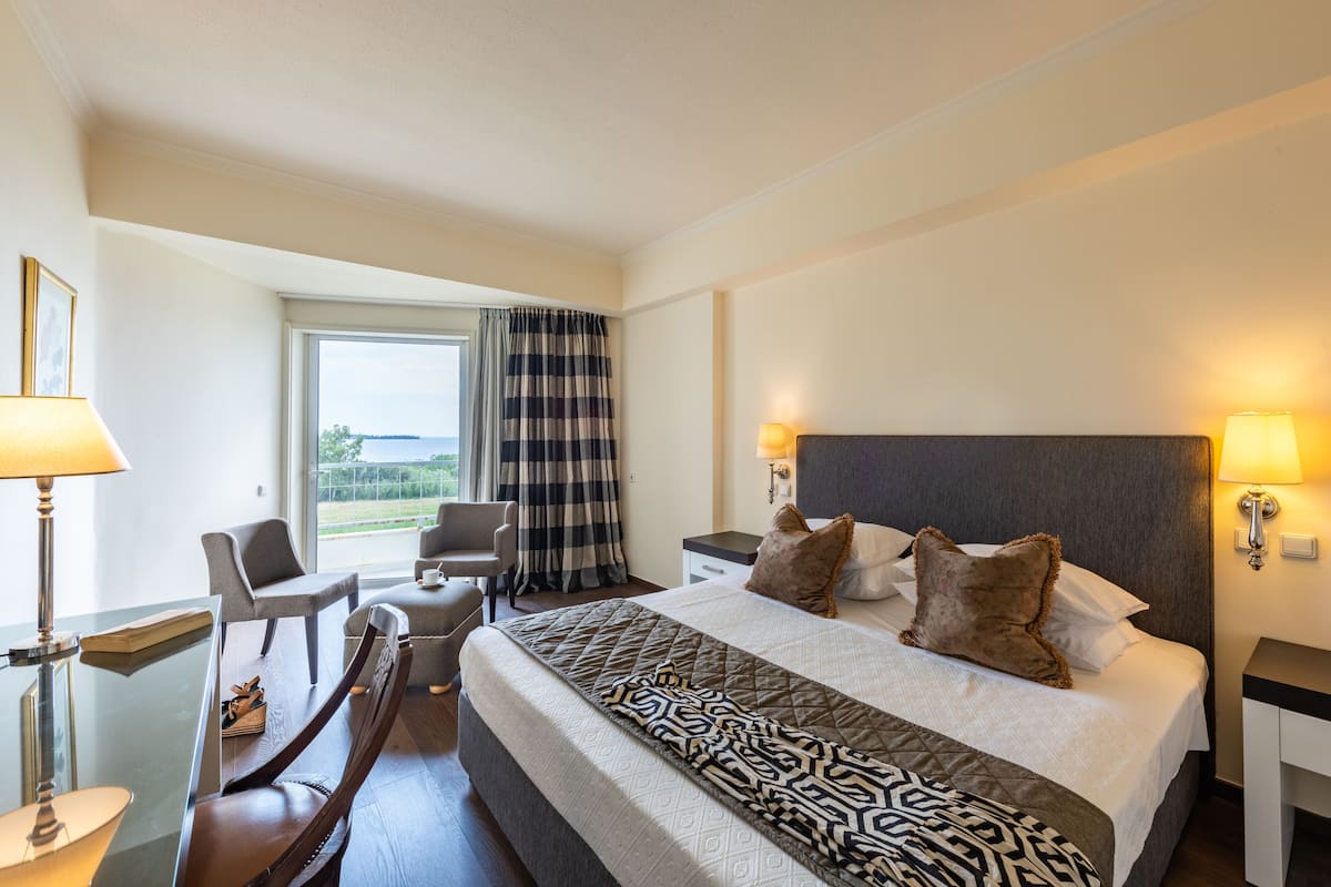 A hotel room with a large bed, a desk with a chair, a window with curtains, and a view of the outdoors.