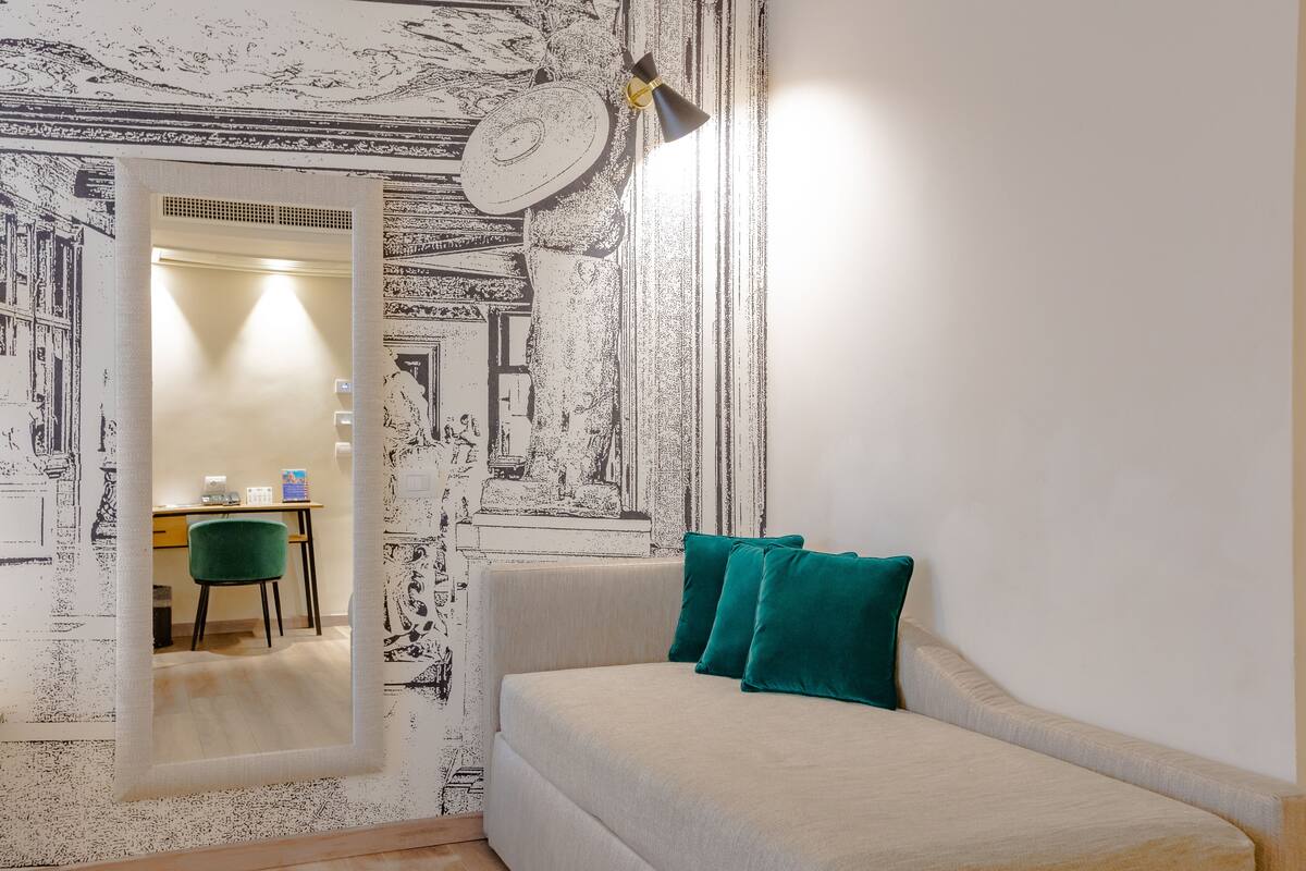 Une pièce comprenant un canapé, deux coussins de couleur turquoise, un bureau avec une chaise et un miroir qui reflète une porte et un luminaire.