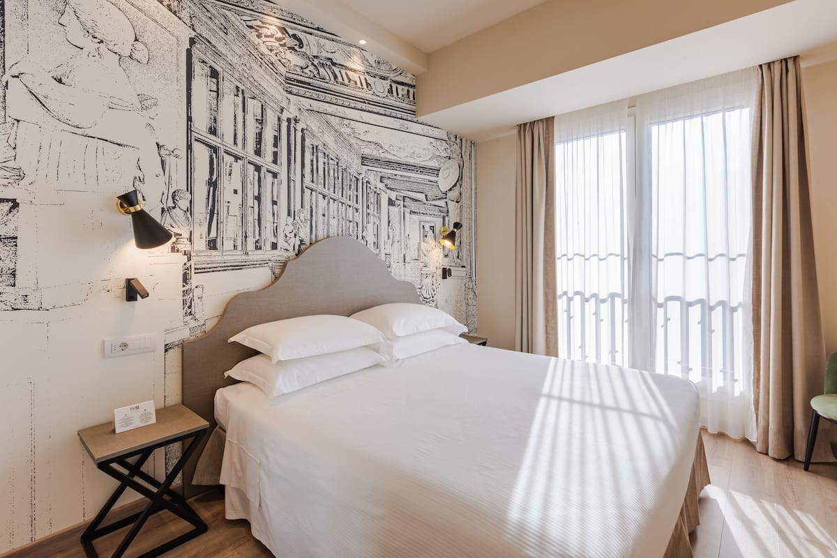 Un lit bien fait, avec une tête de lit, une table de chevet sur laquelle se trouve une lampe, et un mur orné d’une fresque détaillée.