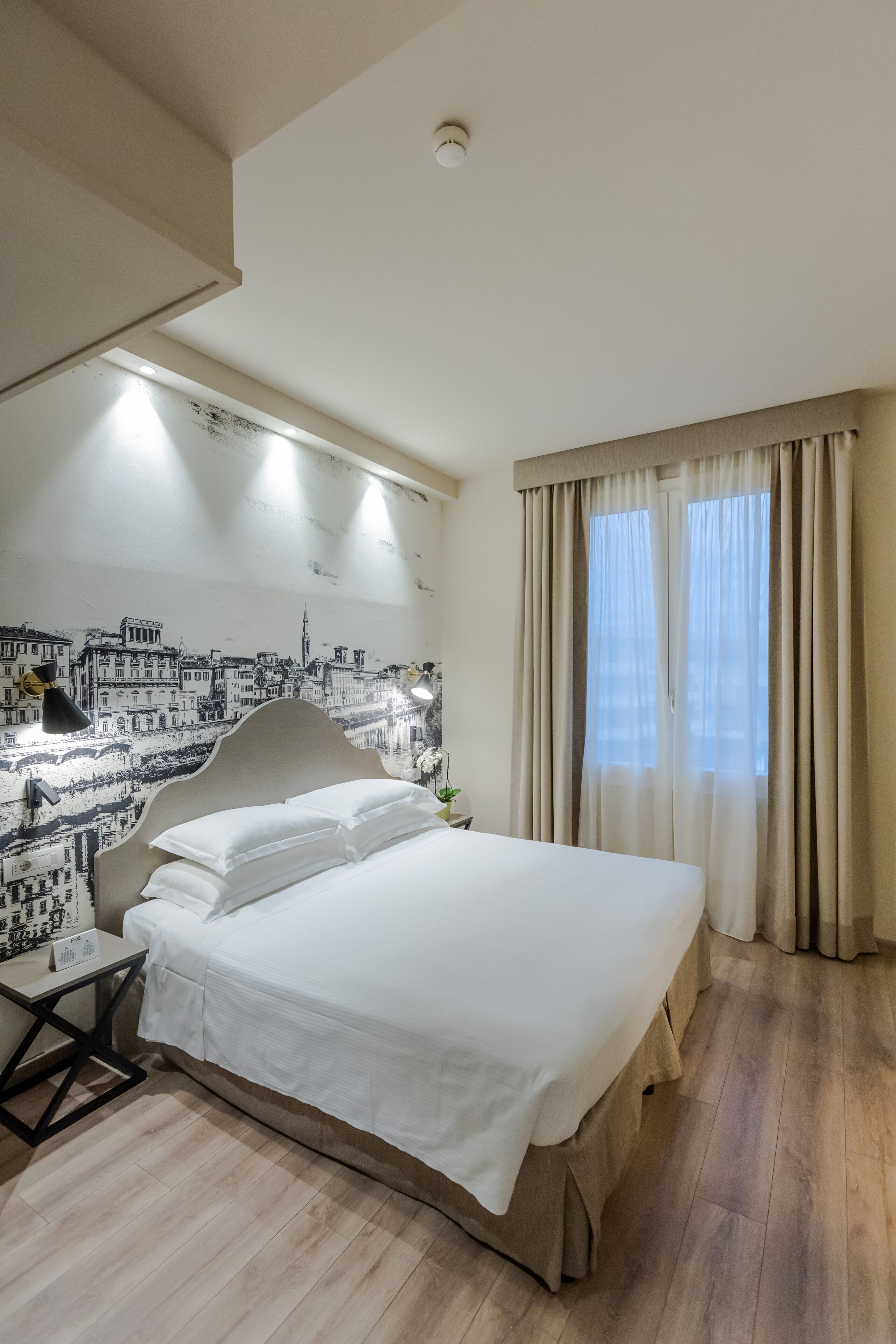 A neatly arranged bedroom with a large bed, a bedside table, a lamp, and a wall with a cityscape mural.