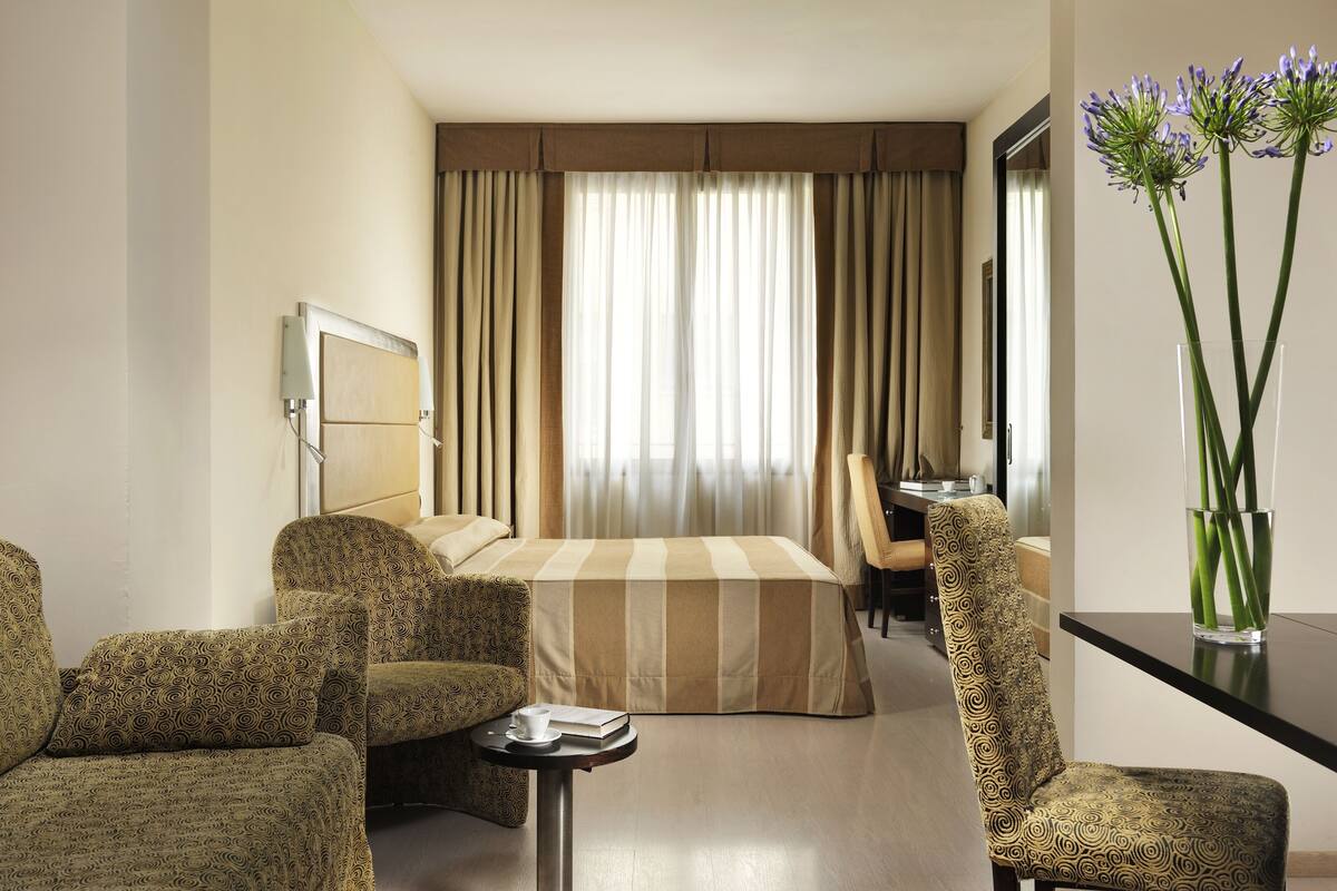 Une chambre d’hôtel avec un lit, deux fauteuils à motifs, une petite table avec un livre et une tasse, un bureau avec une chaise et un vase de fleurs.