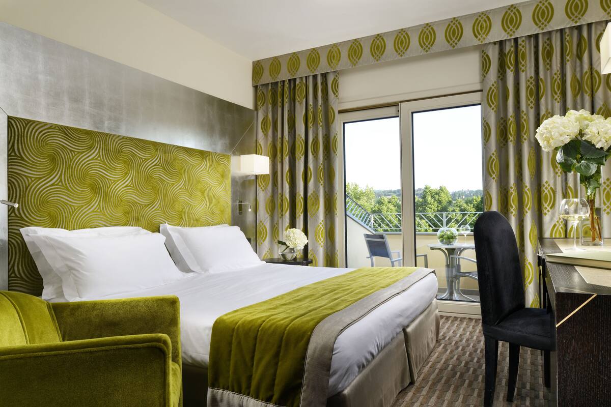 Une chambre d’hôtel avec un grand lit, un bureau et une chaise. Elle dispose d’un balcon avec vue sur la verdure.